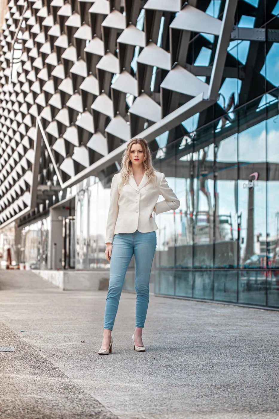 Woman wearing a white blazer, blue jeans and closed shoes outside an office building.