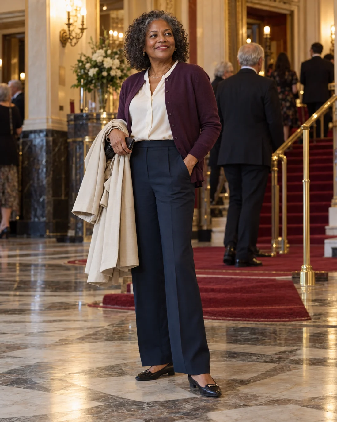 Woman in dark trousers and a light blouse carrying a coat in a theatre foyer.