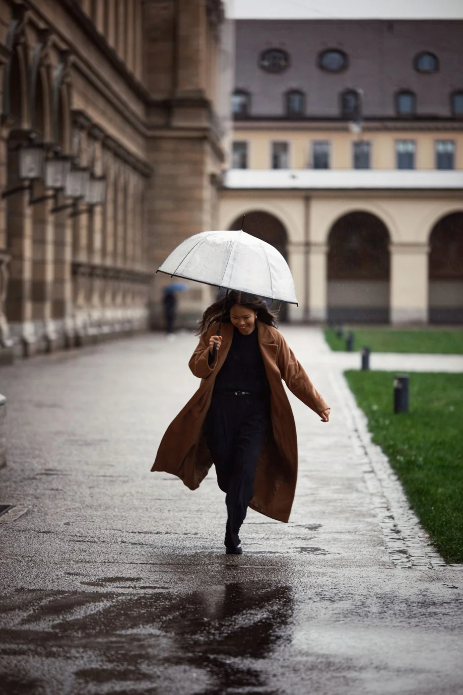 Woman in a brown coat walking through the city with an umbrella.