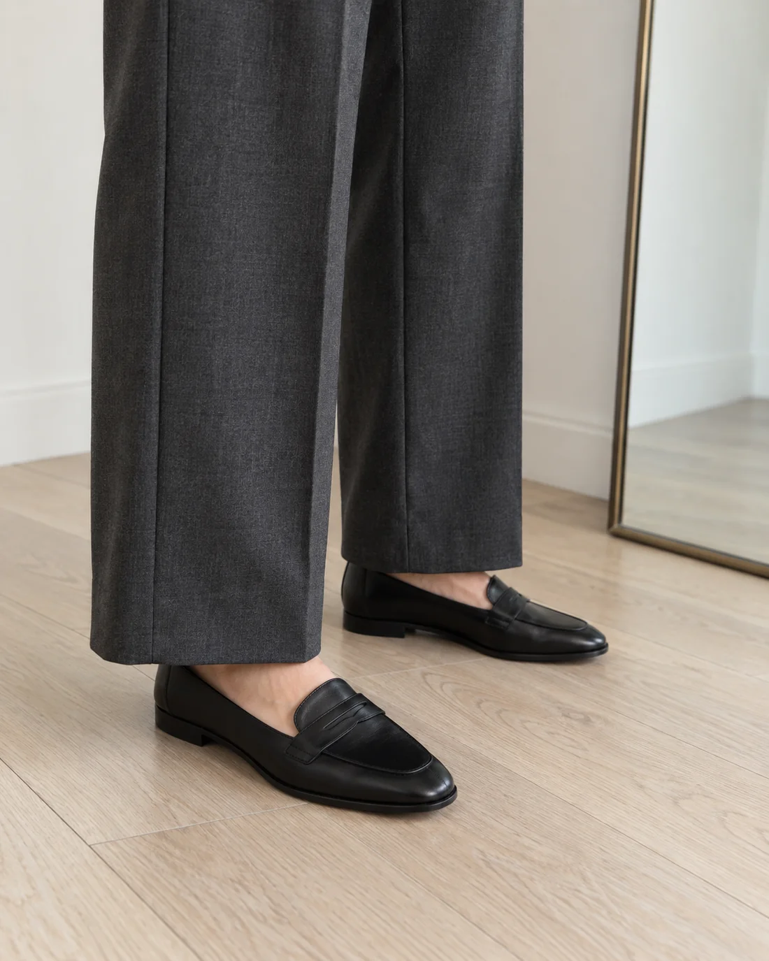 Grey trouser hems above black flat loafers on a wooden floor.