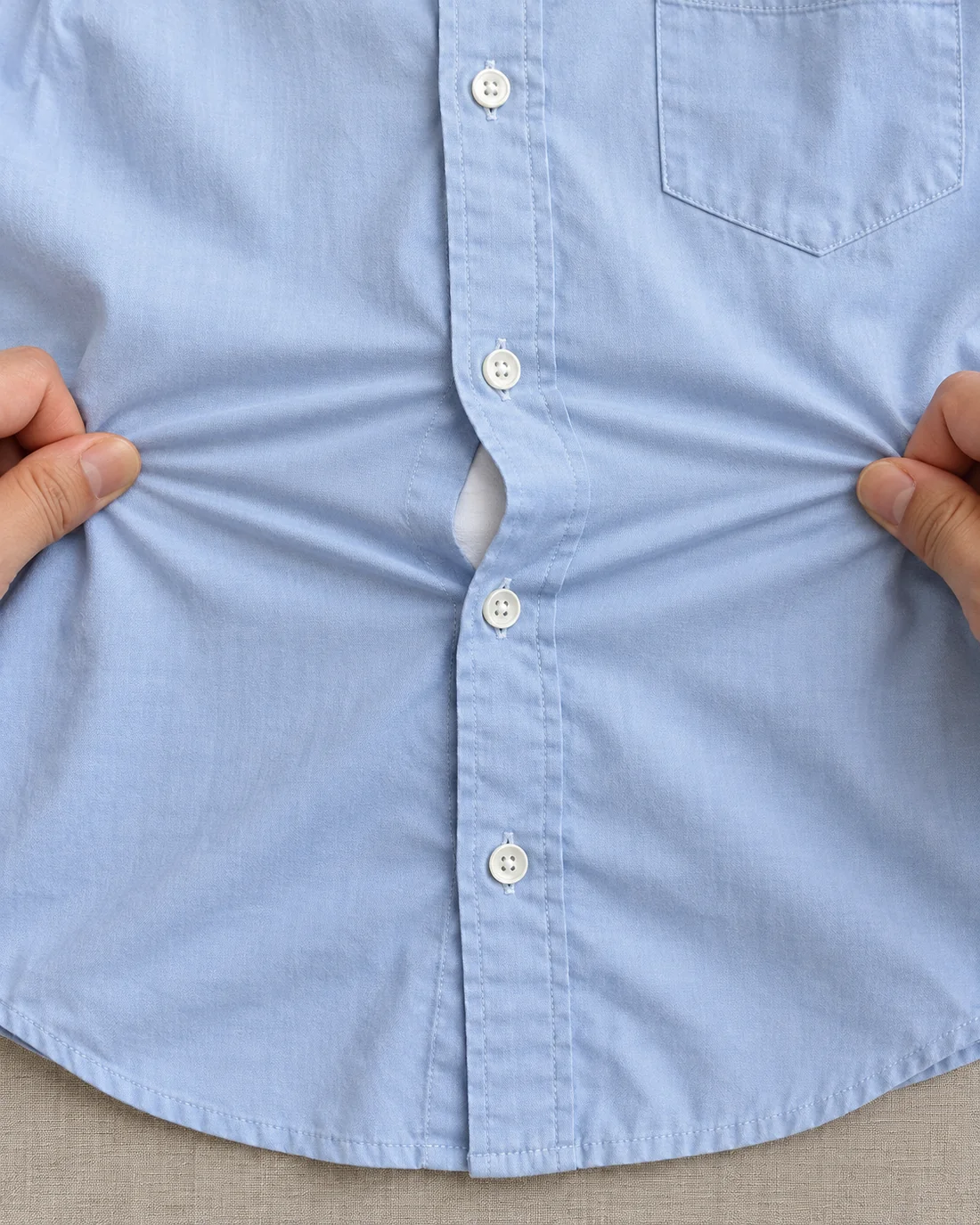 Hands gently pull a flat blue shirt sideways, creating a gap between two buttons.